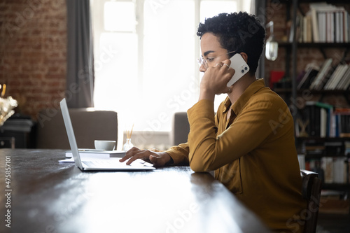 Side view focused young African American businesswoman in eyewear involved in cellphone call conversation working on computer, giving professional consultation to client or negotiating project.