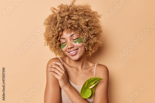 Curly gentle curly haired young woman keeps eyes closed touches shoulder applies green collagen patches to remove dark circles isolated over beige background gets pleasure from skin care routine