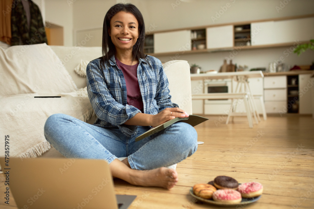 Adorable young woman with dark skin sitting on floor in casual clothes holding notebook, taking notes, using laptop, siting near plate with doughnuts against kitchen interior, leaning on couch