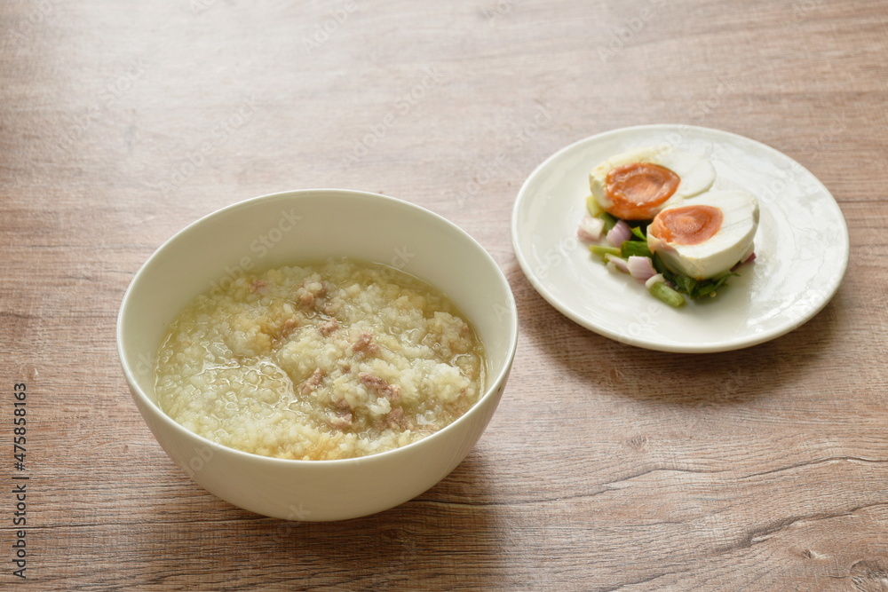 rice porridge with chop pork dressing soy sauce soup in bowl eat couple boiled spicy salty egg half cut topping slice shallot and chili salad on plate rice porridge with chop pork dressing soy sauce soup in bowl eat couple boiled spicy salty egg half cut topping slice shallot and chili salad on plate