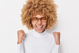 Yes I did it. Overjoyed curly haired young woman makes fist bumps celebrates personal achievements laughs happily with closed eyes wears spectacles and turtleneck isolated over white background