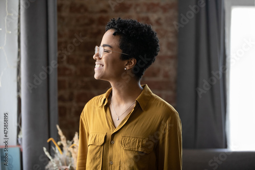 Happy dreamy millennial African American woman in eyeglasses looking in distance, imagining good future, remembering positive life moments, feeling inspired posing in modern loft style home or office.
