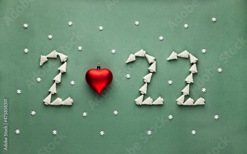 Numbers 2022 from new year tree shaped marshmellow candies and red heart toy on green background with snowflakes.