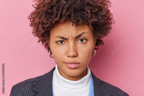 Headshot of displeased young curly haired woman frowns face raises eyebrows dressed in elegant clothes listens something attentively isolated over pink background. Negative human emotions concept