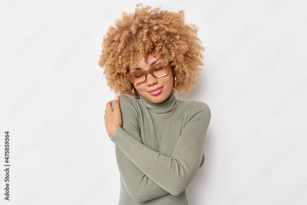 Tender curly haired woman embraces herself wraps body with own hands feels cozy keeps eyes closed wears spectacles and turtleneck isolated over white background. Comfort and romance concept. Tender curly haired woman embraces herself wraps body with own hands feels cozy keeps eyes closed wears spectacles and turtleneck isolated over white background. Comfort and romance concept.