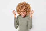 Mad emotional overemotive woman with curly hair screams loudly shakes hands near head keeps mouth opened feels very happy wears spectacles and casual poloneck isolated over white background.