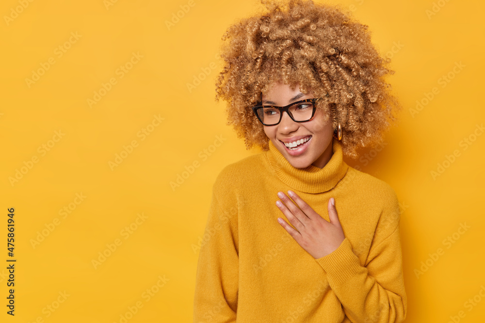 Positive sincere curly woman keeps hand on chest looks away happily smiles broadly laughs at something funny wears spectacles and casual jumper isolated over yellow background. Emotions concept