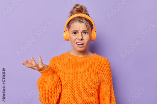Fotografia Puzzled indignant young woman wears casual knitted sweater raises palm feels clueless cannot hear you as listens music in headphones poses against purple background