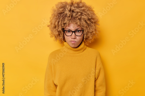 Horizontal shot of outraged curly haired woman frowns face looks angrily being irritated with someone wears transparent glasses and casual sweater isolated over yellow background. Negative emotions © Wayhome Studio