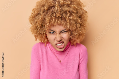 Fotografie, Obraz Photo of irritated young curly haired woman clenches teeth with hateful expression smirks face dressed in casual pink turtleneck isolated over beige background