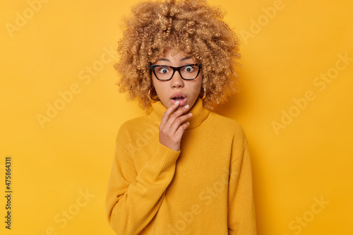 Stunned curly young woman gasps from wonder holds breath stares at something horrible keeps jaw dropped dressed in casual jumper isolated over vivid yellow background. Human reactions concept