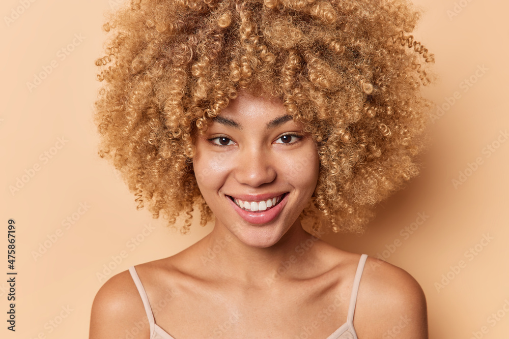 Close up portrait of positive curly haired woman smiles gently has silky healthy skin no make up poses bare shoulders looks directly at camera isolated over beige background. Beauty concept. Close up portrait of positive curly haired woman smiles gently has silky healthy skin no make up poses bare shoulders looks directly at camera isolated over beige background. Beauty concept.
