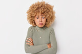 Puzzled offended woman keeps arms folded looks disappointed dressed in casual turtleneck has curly hair feels disappointed and embrassed isolated over white background. Negative face expressions