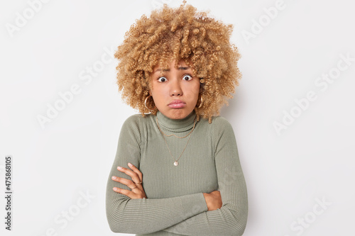 Puzzled offended woman keeps arms folded looks disappointed dressed in casual turtleneck has curly hair feels disappointed and embrassed isolated over white background. Negative face expressions photo