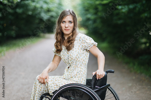 Portrait of young caucasian woman looking at camera while sitting in wheelchair in stylish summer dress. Leisure activity at green park. Lifestyles of people with disability.