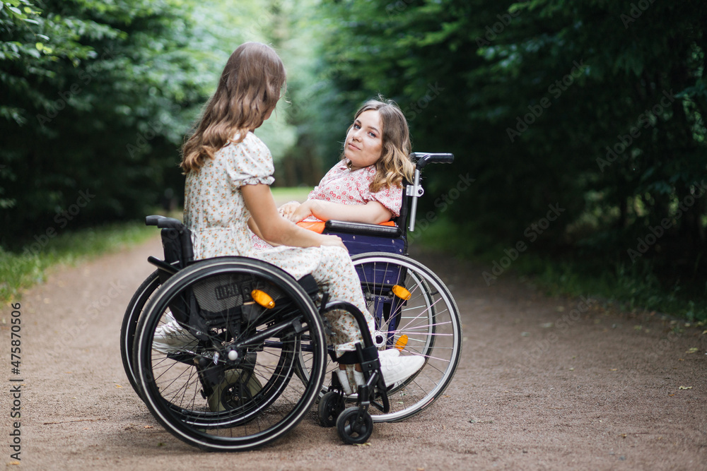Two disabled women in stylish dressed enjoying summer walk at green park. Young sitting in wheelchair and looking at camera. Friendship concept.