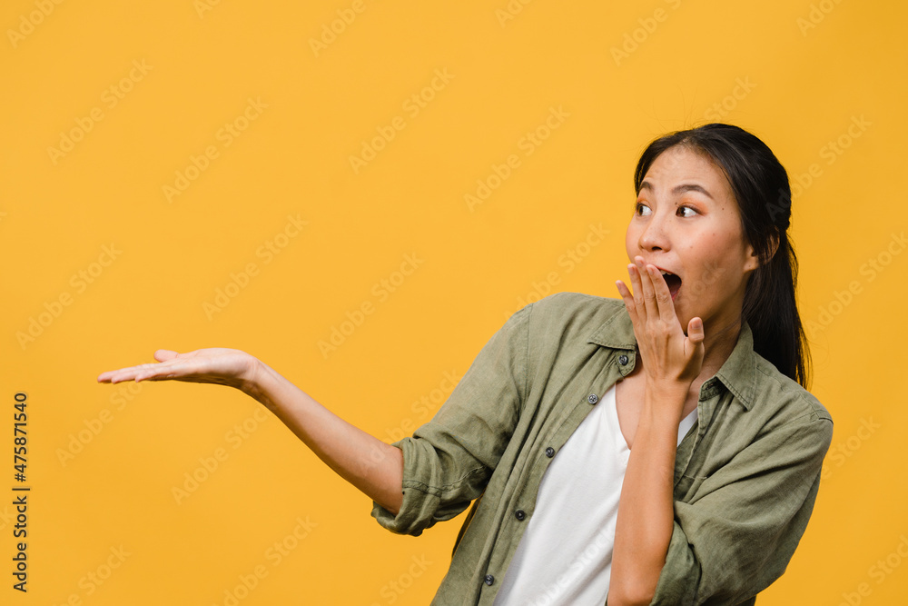 Portrait of young Asian lady smiling with cheerful expression, shows something amazing at blank space in casual clothing and standing isolated over yellow background. Facial expression concept. Portrait of young Asian lady smiling with cheerful expression, shows something amazing at blank space in casual clothing and standing isolated over yellow background. Facial expression concept.