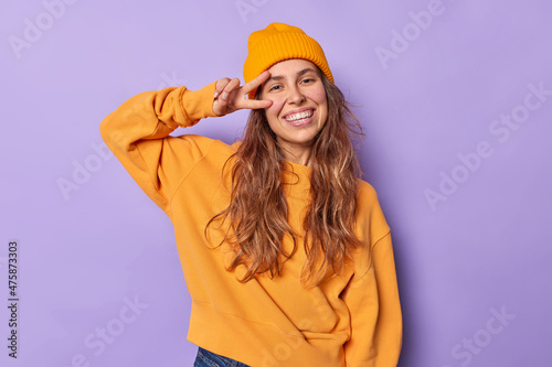 Pretty positive young woman makes peace gesture over eye feels glad enjoys life wears casual yellow jumper and hat isolated over purple background salutes you with happy expression makes v sign