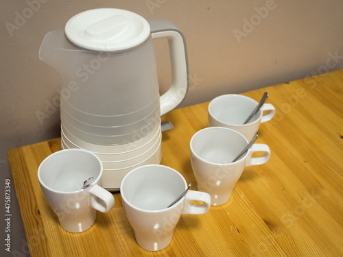White electric kettle and four white ceramic mugs with teaspoons on wooden table