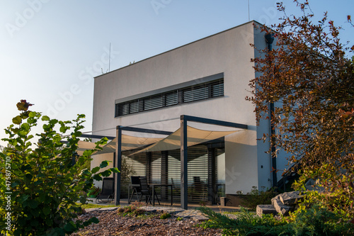 Modern, white, cube, elegant, minimalist style passive house with large panoramic windows, grey shutters in maintained garden in sunset warm light. Wooden terrace. Garden furniture. photo