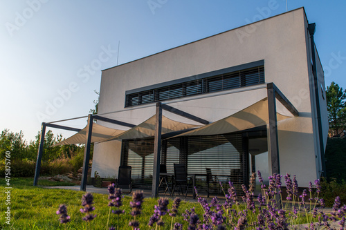 Modern, white, cube, elegant, minimalist style passive house with large panoramic windows, grey shutters in maintained garden. Wooden terrace. Sunny day. Lavender in front.