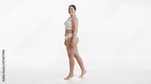 Horizontal long shot of young pretty Caucasian plus size woman in white underwear stands on tiptoes and puts her hand on her waist smiling wide to the camera on white background | Body care concept photo