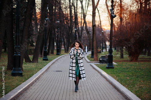 Full length portrait of fashionable elegant woman in checkered black and white coat an green leather dress walking in the park outdoors in autumn or winter.
