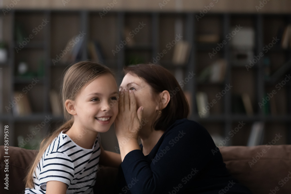 In on a little secret. Older grandmother sit on cozy sofa close to smiling preteen girl granddaughter whisper funny rumor on child ear. Excited tween kid listen to cute gossip telling by mature granny In on a little secret. Older grandmother sit on cozy sofa close to smiling preteen girl granddaughter whisper funny rumor on child ear. Excited tween kid listen to cute gossip telling by mature granny