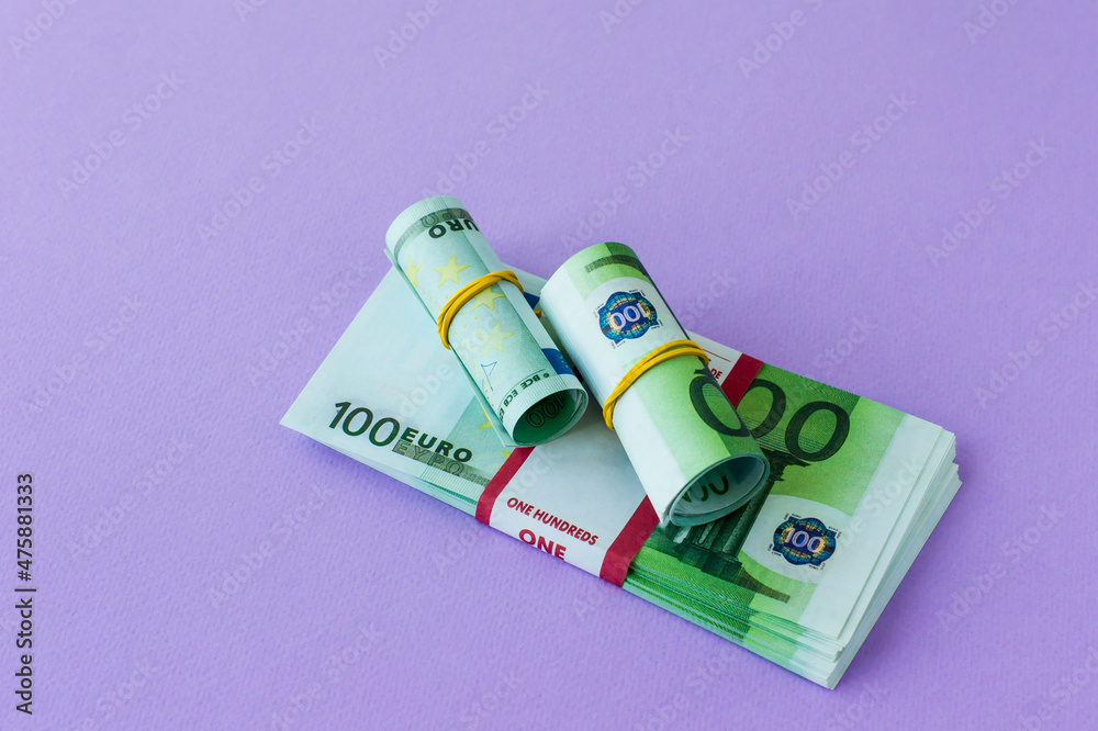 a pack of 100 euro banknotes in a bank package and two rolls under an elastic band on it. lilac background. a pack of 100 euro banknotes in a bank package and two rolls under an elastic band on it. lilac background.