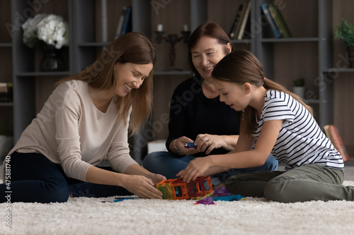 Leisure time. Happy tween girl young mother old age grandmother spend free time on carpet create fantasy figures from magnetic constructor. Friendly family of 3 diverse females enjoy playing together