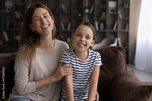 Having fun together with mommy. Overjoyed preteen age daughter and adult mother sit on couch at living room laugh joke look at camera. Portrait of joyful little girl spend funny time with young nanny