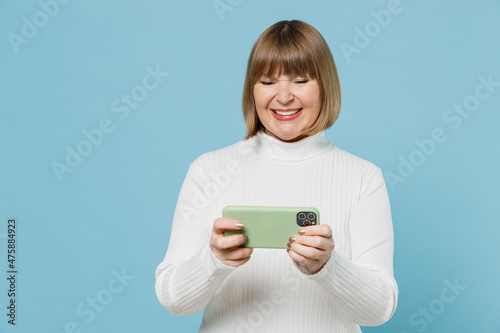 Elderly fun woman 50s in white knitted sweater using play racing app on mobile cell phone hold gadget smartphone for pc video games isolated on plain blue color background . People lifestyle concept.