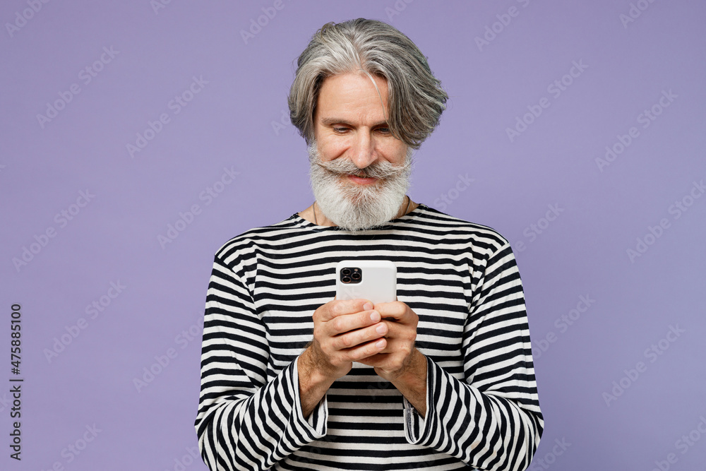 Elderly gray-haired mustache bearded man 50s wearing striped turtleneck hold in hand use mobile cell phone isolated on plain pastel light purple background studio portrait. People lifestyle concept. Elderly gray-haired mustache bearded man 50s wearing striped turtleneck hold in hand use mobile cell phone isolated on plain pastel light purple background studio portrait. People lifestyle concept.