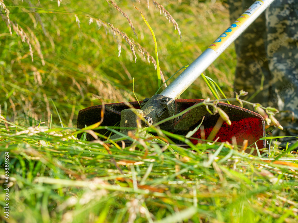 Close-up of a grass trimmer with a nozzle in the form of a metal knife. Care of the plot, cutting grass, tall grass, weeds. View from below from the ground Close-up of a grass trimmer with a nozzle in the form of a metal knife. Care of the plot, cutting grass, tall grass, weeds. View from below from the ground
