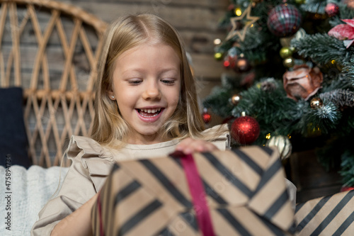 Happy girl sitting by a Christmas tree unwrapping a present