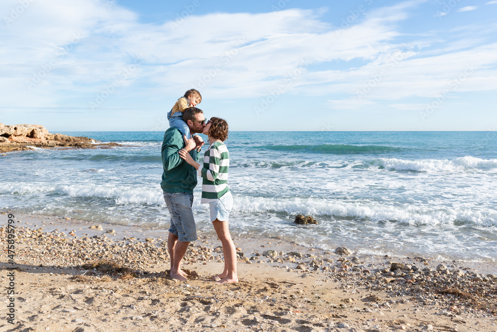 Couple kissing with their baby girl on her father's shoulders at the sea shore. Love and togetherness. 