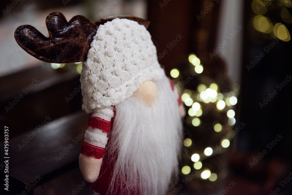 Ozdoba świąteczna, skrzat w ciepłej czapce, z białą brodą i porożem renifera. W tle światełka choinkowe - obrazy, fototapety, plakaty 