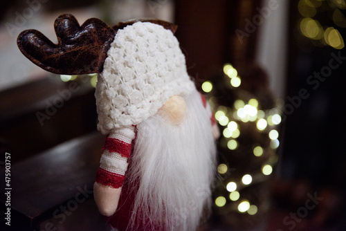 Ozdoba świąteczna, skrzat w ciepłej czapce, z białą brodą i porożem renifera. W tle światełka choinkowe