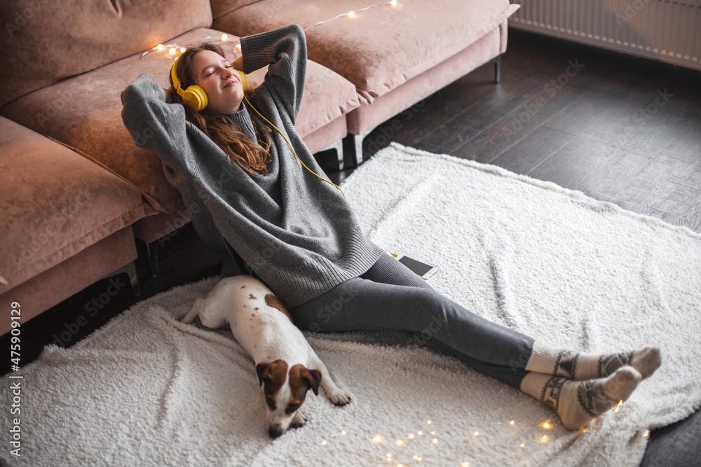 Obraz premium Cheerful young woman listening to music in headphones sitting near the sofa in the room
