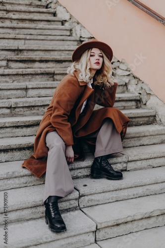 Beautiful girl with blonde hair sits on the stairs in the old city on the street in brown trousers, a brown coat, a white blouse, black shorts and a hat