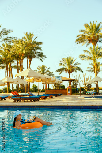 A woman in a sun hat in the pool in a swimming circle with a laptop on the background of palm trees and sun loungers.