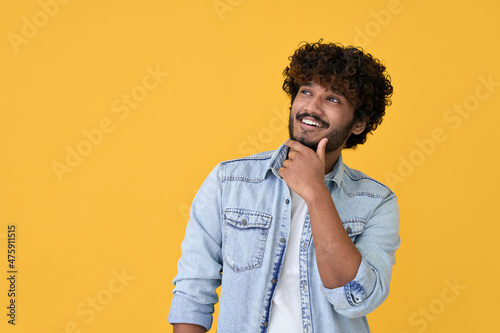 Smiling curious young indian man holding hand on chin looking interested aside at copy space isolated on yellow background thinking of shopping opportunities, planning purchase or dreaming concept. photo