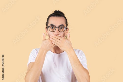 handsome latin man with glasses to see, covers his mouth face of surprise or fright, beige background