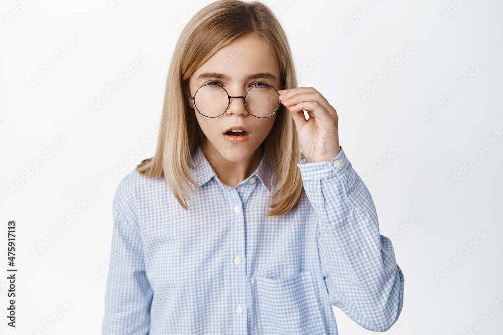 Portrait of blond girl, little kid in glasses squinting, has difficulties reading without eyewear, standing over white background