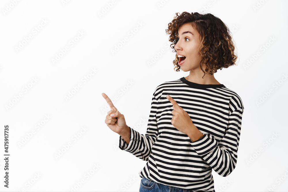 Stylish modern girl with short curly hair, showing advertisement on copy space, pointing left at empty side and smiling, standing over white background Stylish modern girl with short curly hair, showing advertisement on copy space, pointing left at empty side and smiling, standing over white background