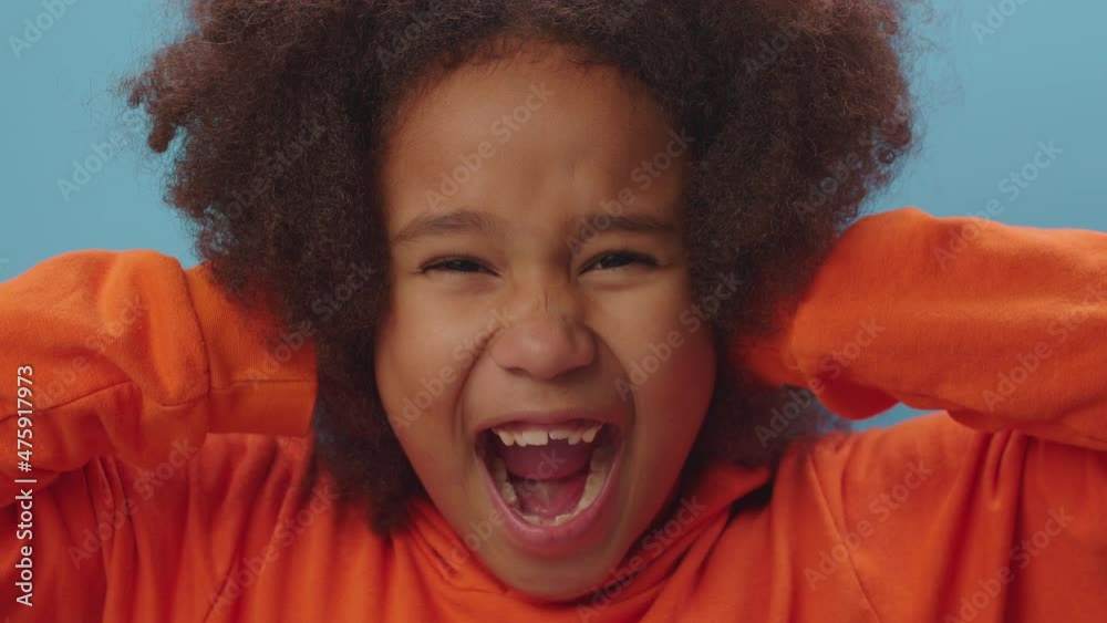 Close up of pretty African American girl closes her ears with hands and screaming 'no' standing on blue background. Kid doesn't want to hear anything. Child's protest. Close up of pretty African American girl closes her ears with hands and screaming 'no' standing on blue background. Kid doesn't want to hear anything. Child's protest.