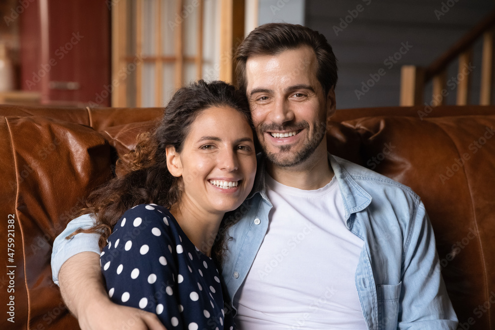 Portrait of smiling loving bonding young family couple resting on sofa, happy millennial man cuddling beautiful hispanic woman enjoying lovely sweet tender relaxed weekend pastime together at home.