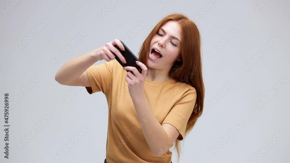 Gambling. young girl 20s in a yellow t-shirt posing using a mobile phone, holding a gadget, addicted to video computer games, isolated on a gray background Gambling. young girl 20s in a yellow t-shirt posing using a mobile phone, holding a gadget, addicted to video computer games, isolated on a gray background