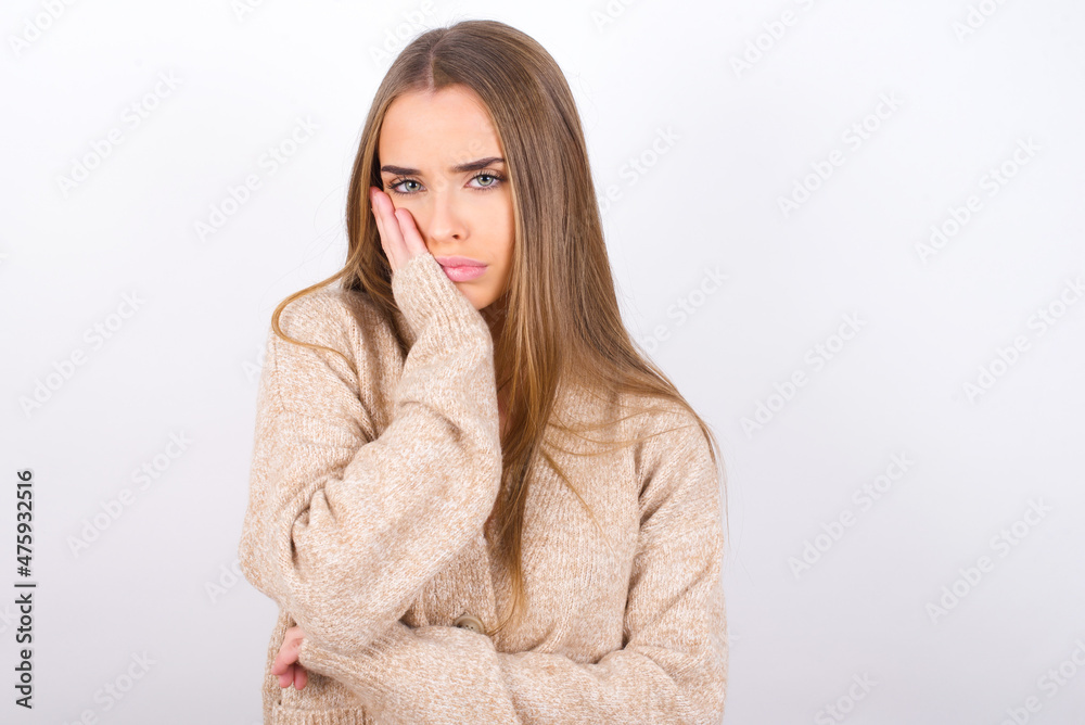Very bored young caucasian girl wearing knitted sweater over white background holding hand on cheek while support it with another crossed hand, looking tired and sick, Very bored young caucasian girl wearing knitted sweater over white background holding hand on cheek while support it with another crossed hand, looking tired and sick,