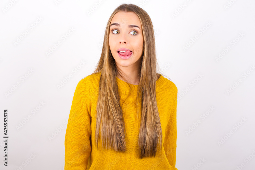 Funny Young caucasian girl wearing yellow sweater over white background makes grimace and crosses eyes plays fool has fun alone sticks out tongue. Funny Young caucasian girl wearing yellow sweater over white background makes grimace and crosses eyes plays fool has fun alone sticks out tongue.